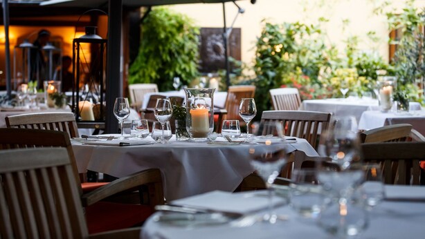 Un área de comedor al aire libre con mesas dispuestas para dos, con manteles blancos, velas y copas de vidrio transparente.
