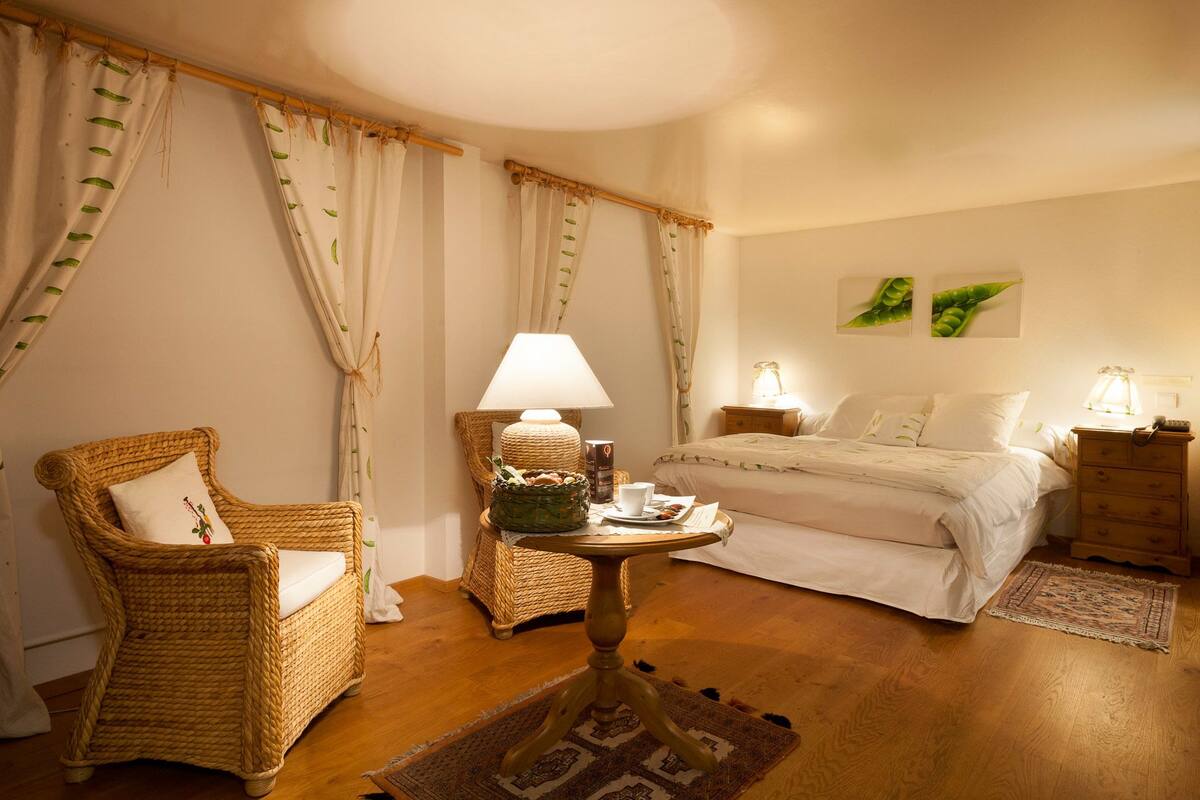 A cozy hotel room with a bed, two wicker chairs, a small round table with a lamp, and a nightstand with a lamp.