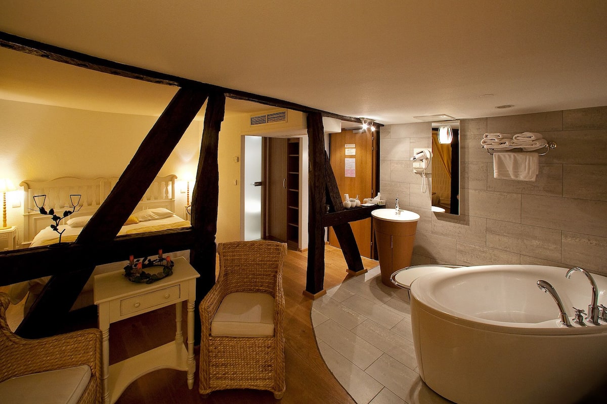 A hotel bathroom with a large bathtub, a sink, a mirror, and a wicker chair.