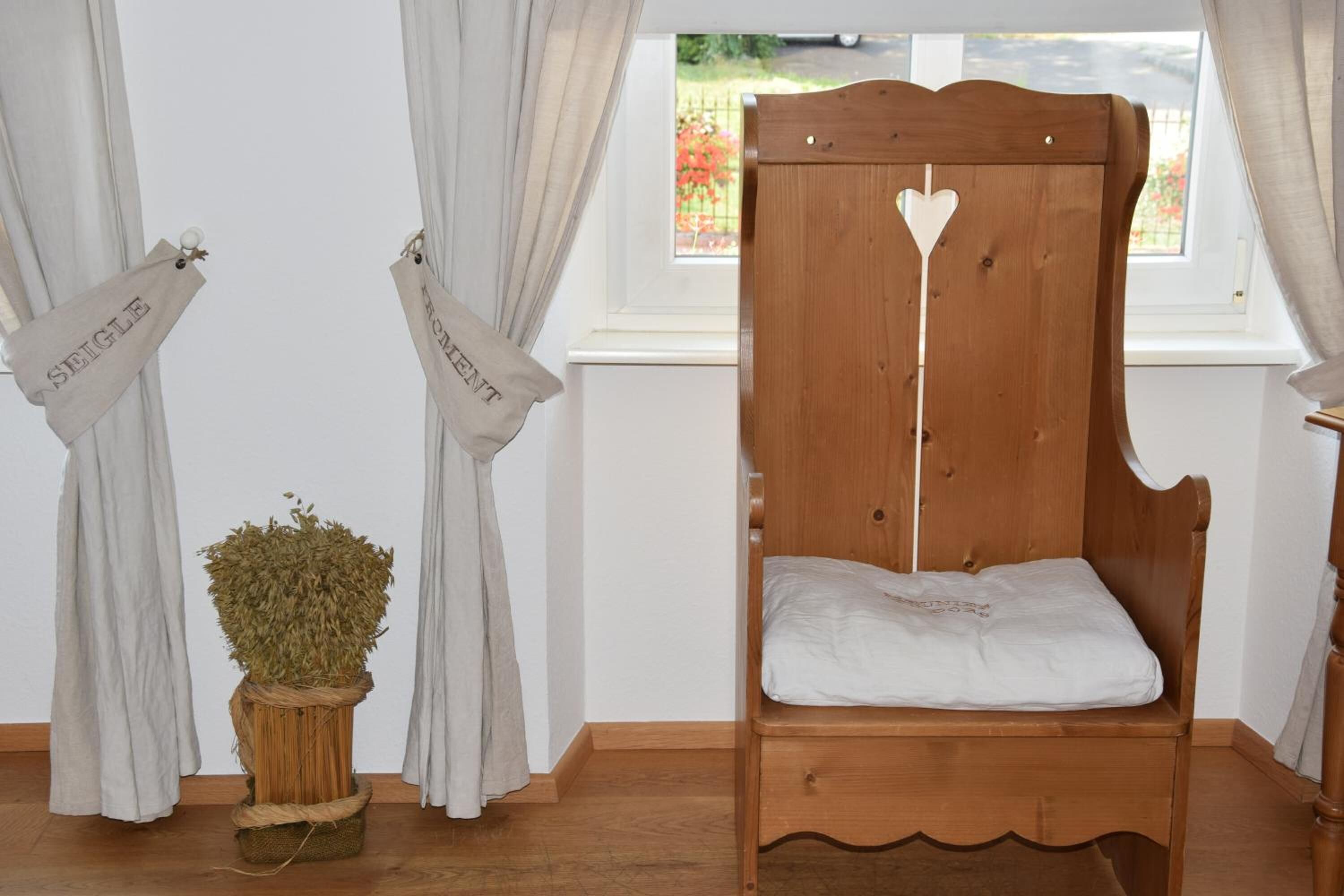 Un lit en bois avec un matelas blanc, dans une pièce où les rideaux sont blancs et où une fenêtre offre une vue sur la verdure.