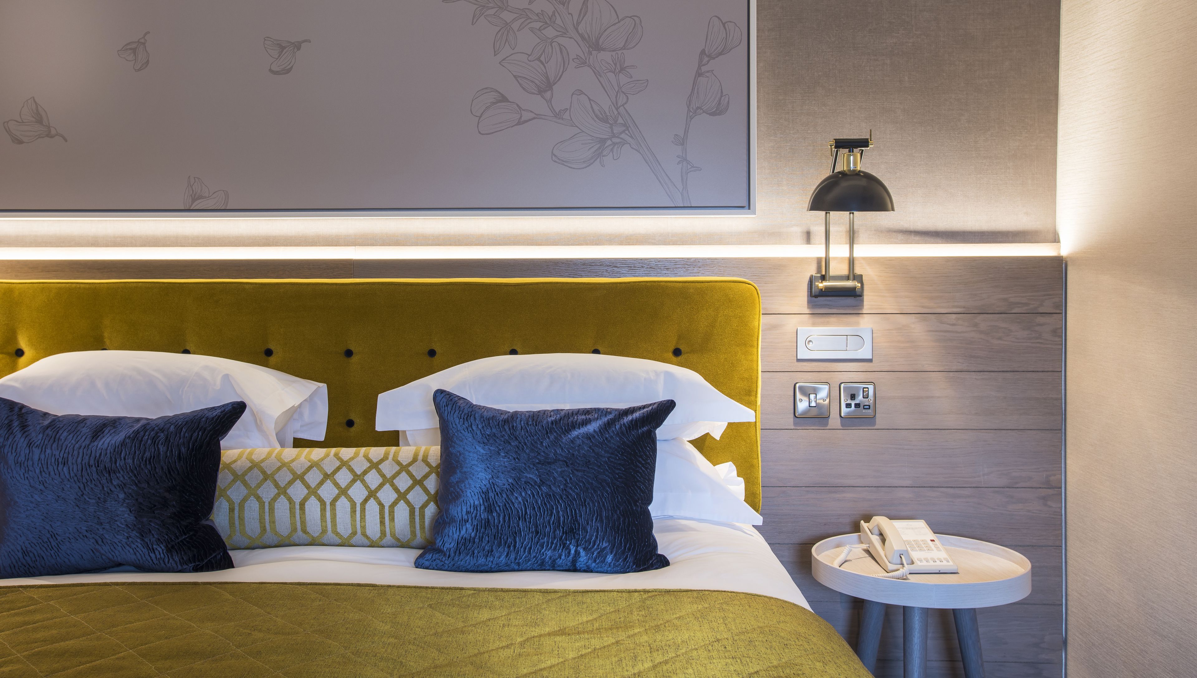A hotel room with a bed, two pillows, a bedside table with a phone, and a wall-mounted lamp.