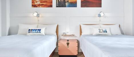 A hotel room with two beds, each with a pillow labeled "BREEZE" and "SALT." There are wall-mounted lamps, a wooden nightstand, and a suitcase.