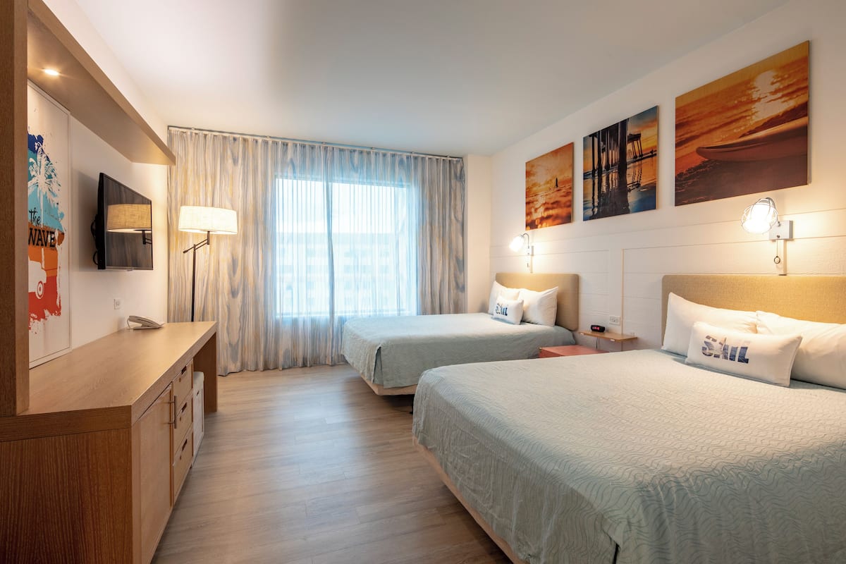 Ein Hotelzimmer mit zwei Betten, einem Schreibtisch, einem Fernseher und Bildern an den Wänden.