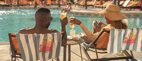 A couple sitting at a poolside table with drinks, surrounded by lounge chairs and umbrellas.