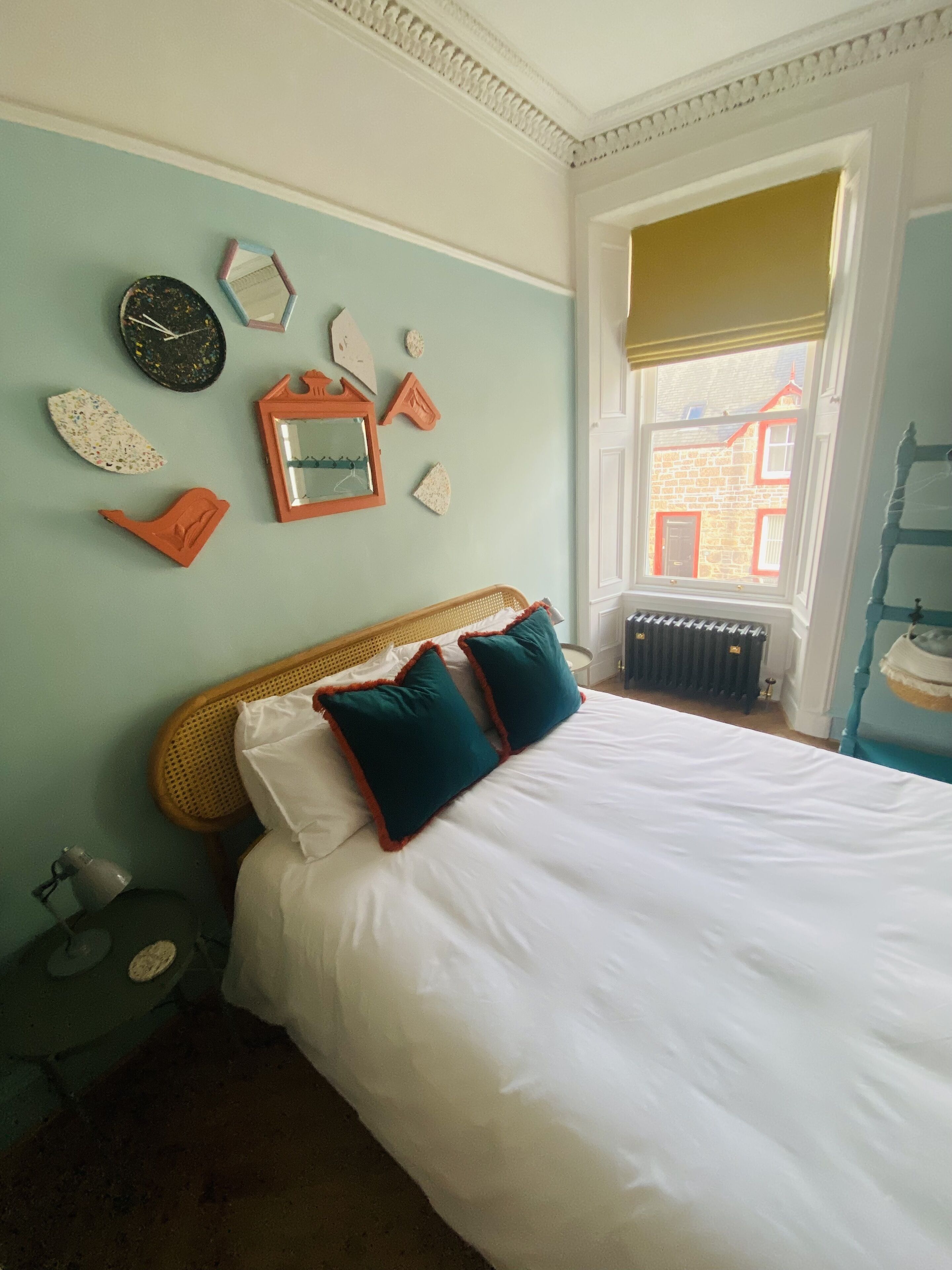 Una cama bien hecha con sábanas blancas y almohadas azul verdosas, un cabecero de madera y una pared decorada con varios objetos, incluyendo un reloj y un espejo.