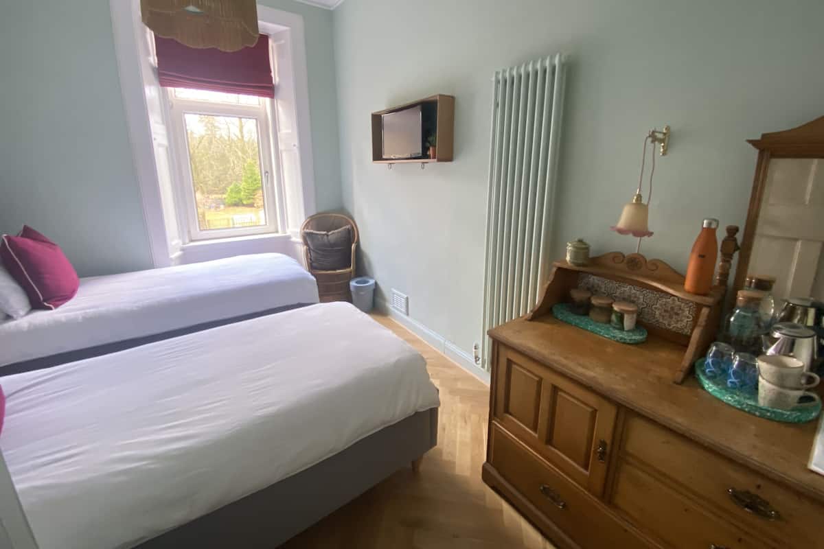 A bedroom with two beds, a dresser, a window with curtains, a radiator, and a small television on the wall.