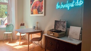 A room with a wooden table, two chairs, a neon sign reading "the breakfast club", a world map suitcase, and a framed sketch on a cabinet. - Ben A'an House (Callander)