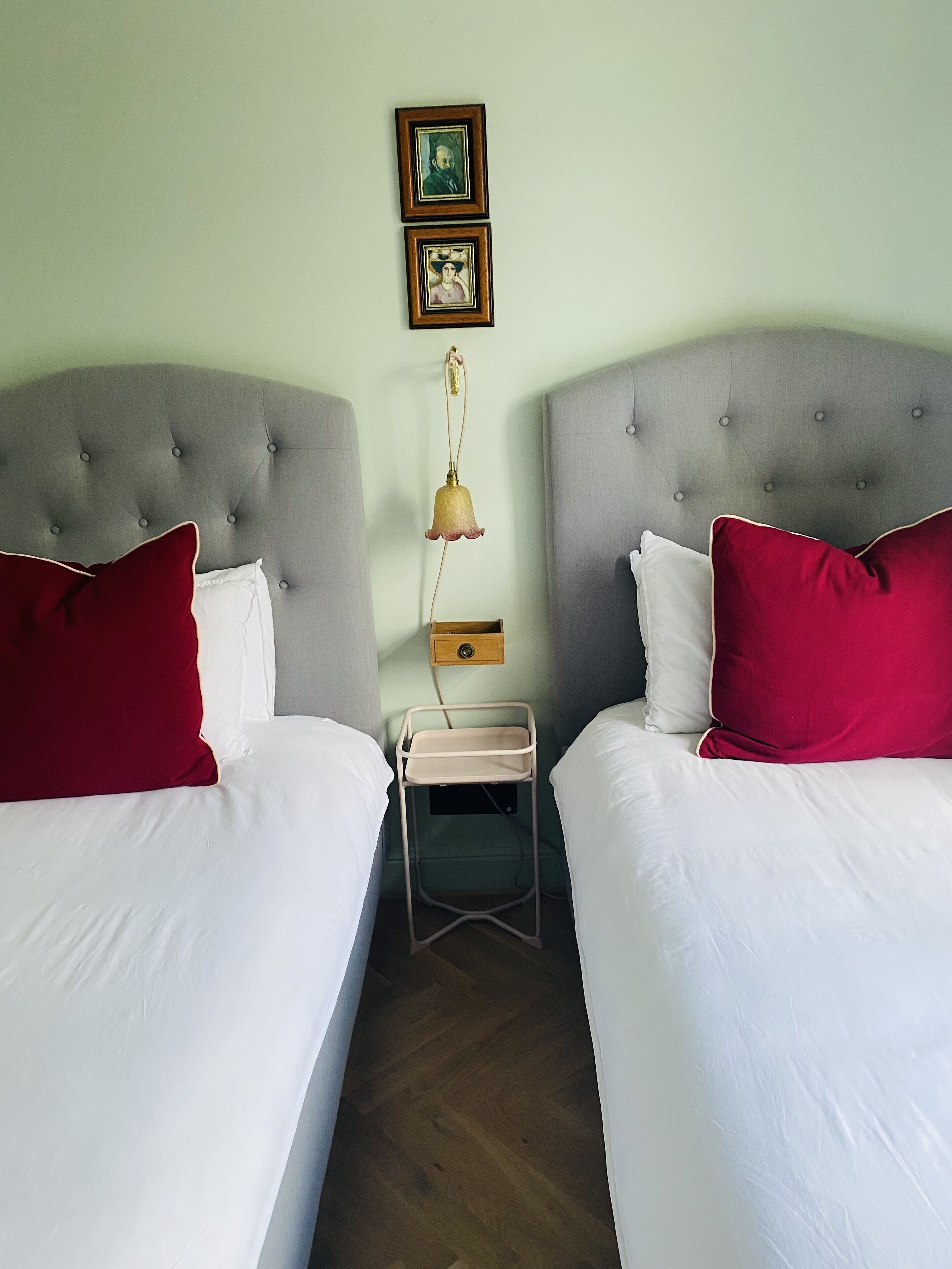 A twin room with two beds, each with a red pillow and white bedding. There is a small bedside table with a lamp and a picture frame on the wall above.