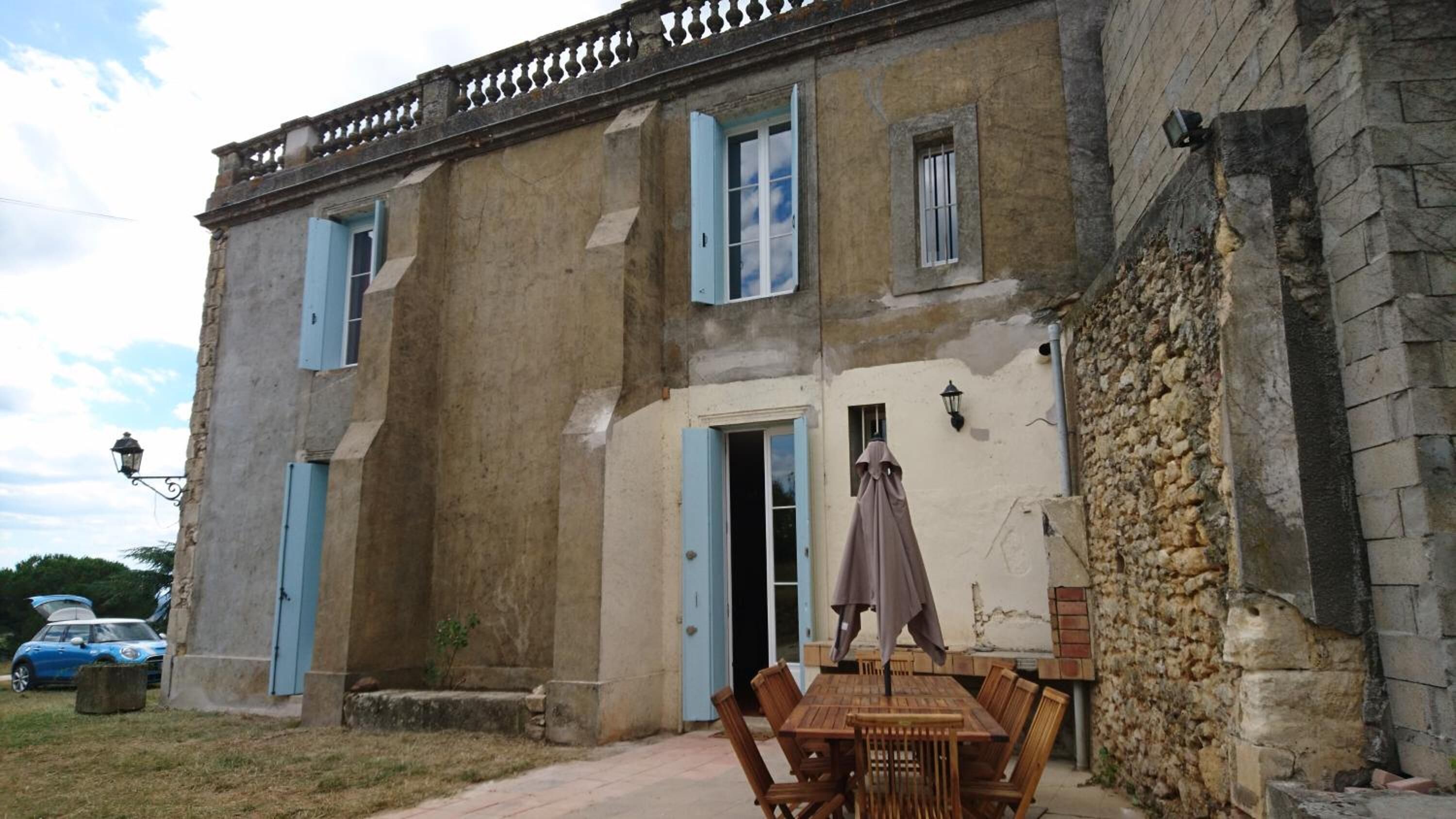 A rustic building with a stone wall, a wooden table with chairs, and a blue door.