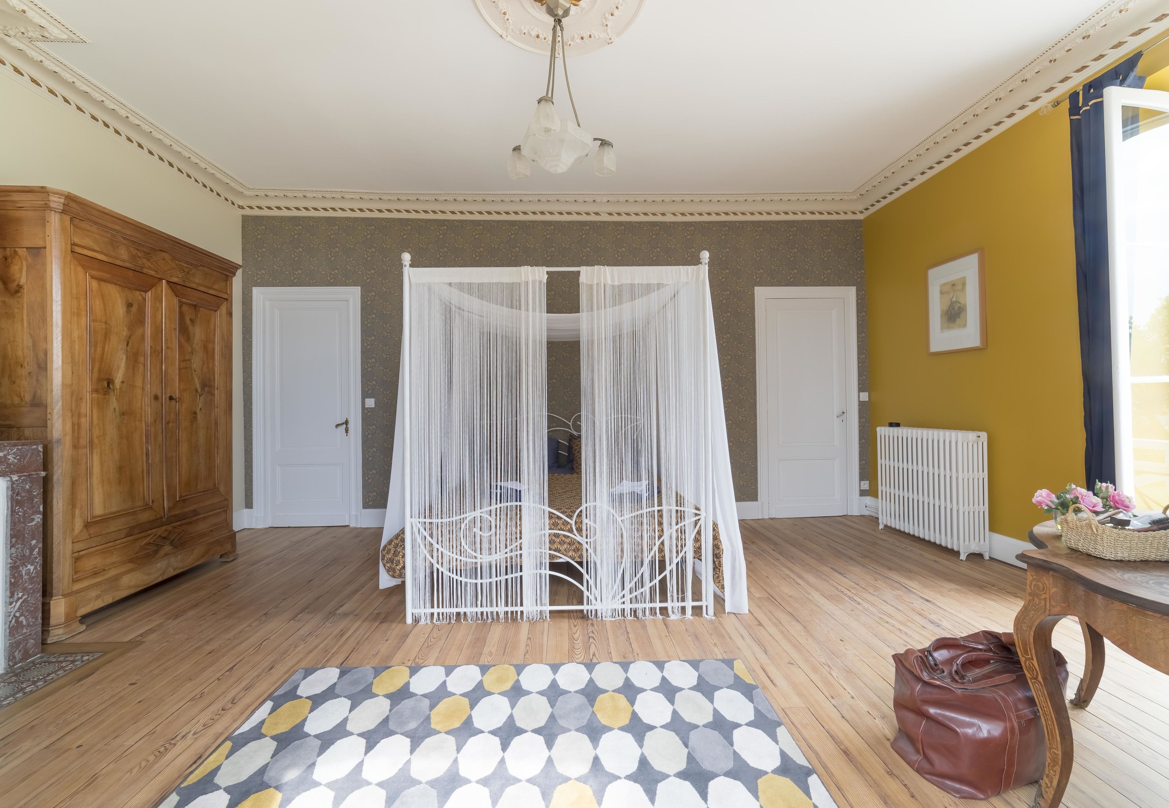Quarto com piso de madeira, tapete estampado, cama com dossel branco, guarda-roupa de madeira e radiador.