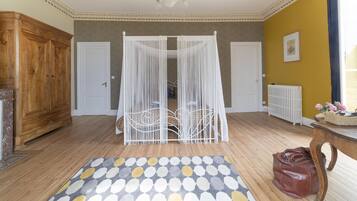 Quarto com piso de madeira, tapete estampado, cama com dossel branco, guarda-roupa de madeira e radiador.