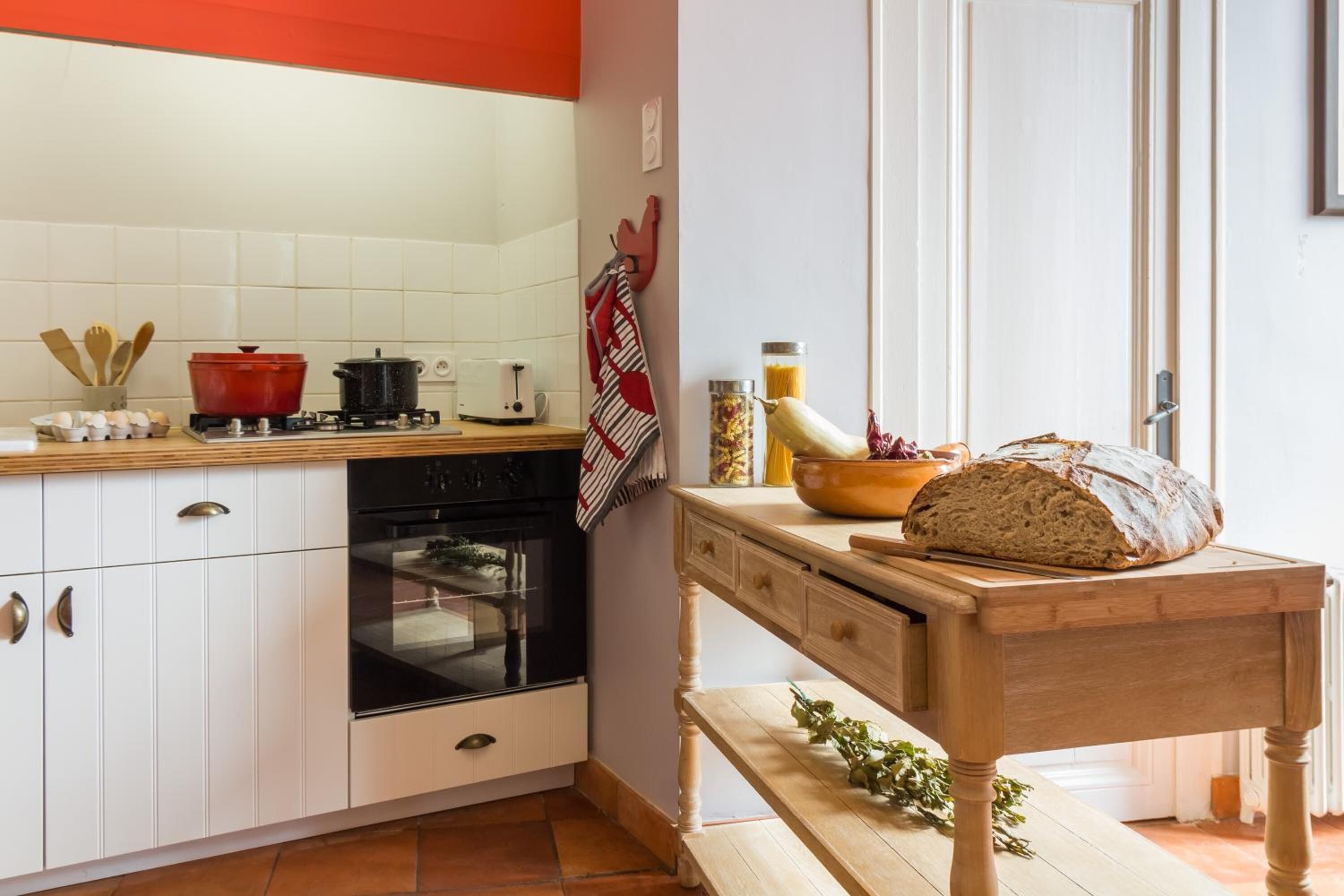 A kitchen with white cabinets, a red pot, a black oven, and a wooden table with bread.