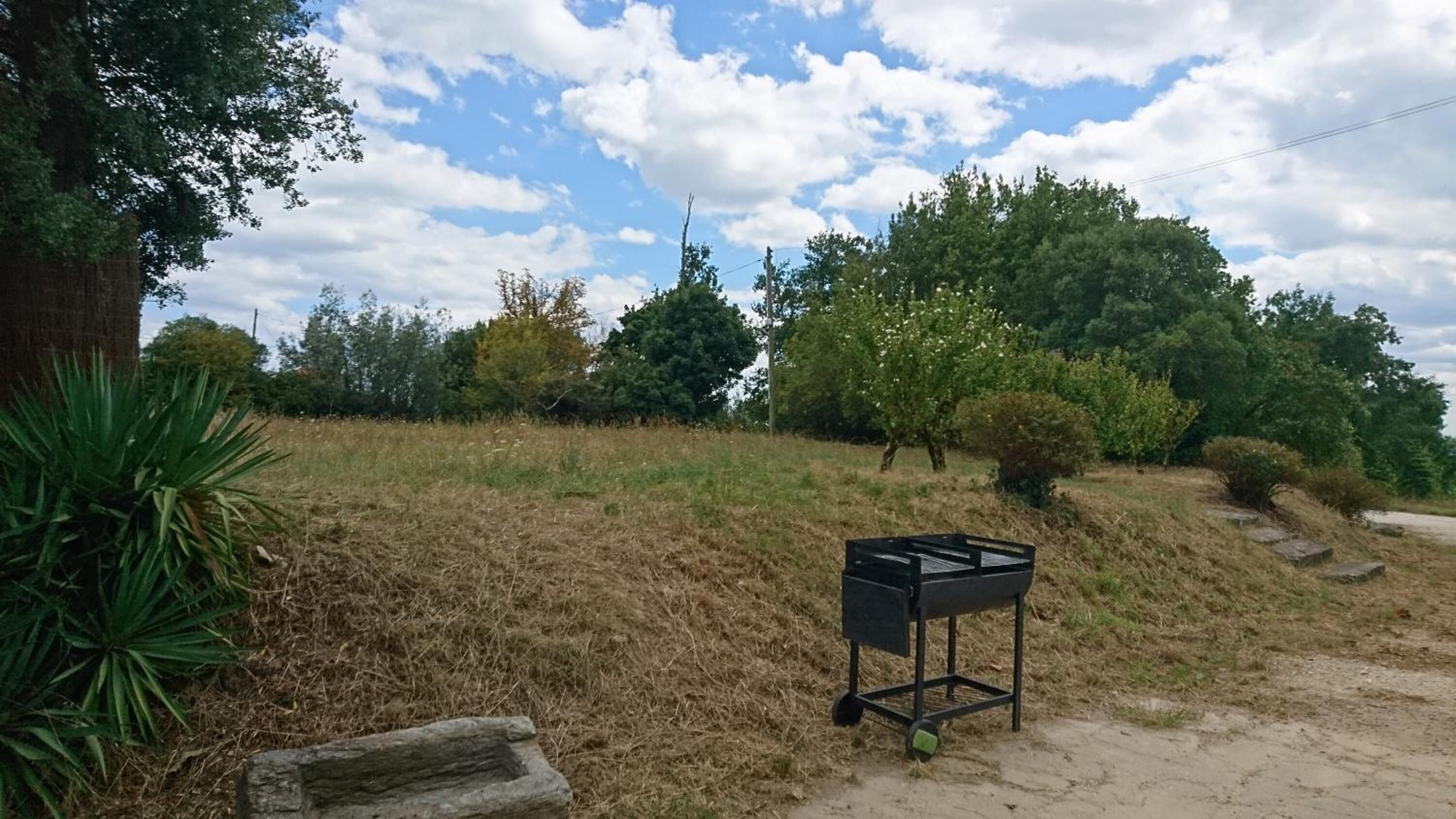 A black grill on a metal stand in a grassy area with trees and a clear sky.