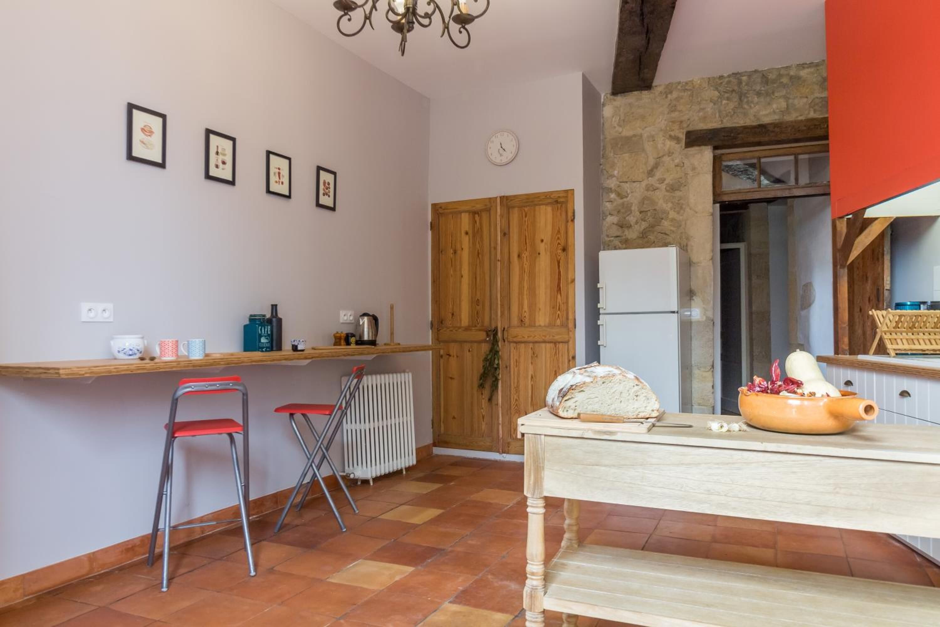 A kitchen with a wooden door, a white refrigerator, a table with bread and a bowl, and two red chairs.