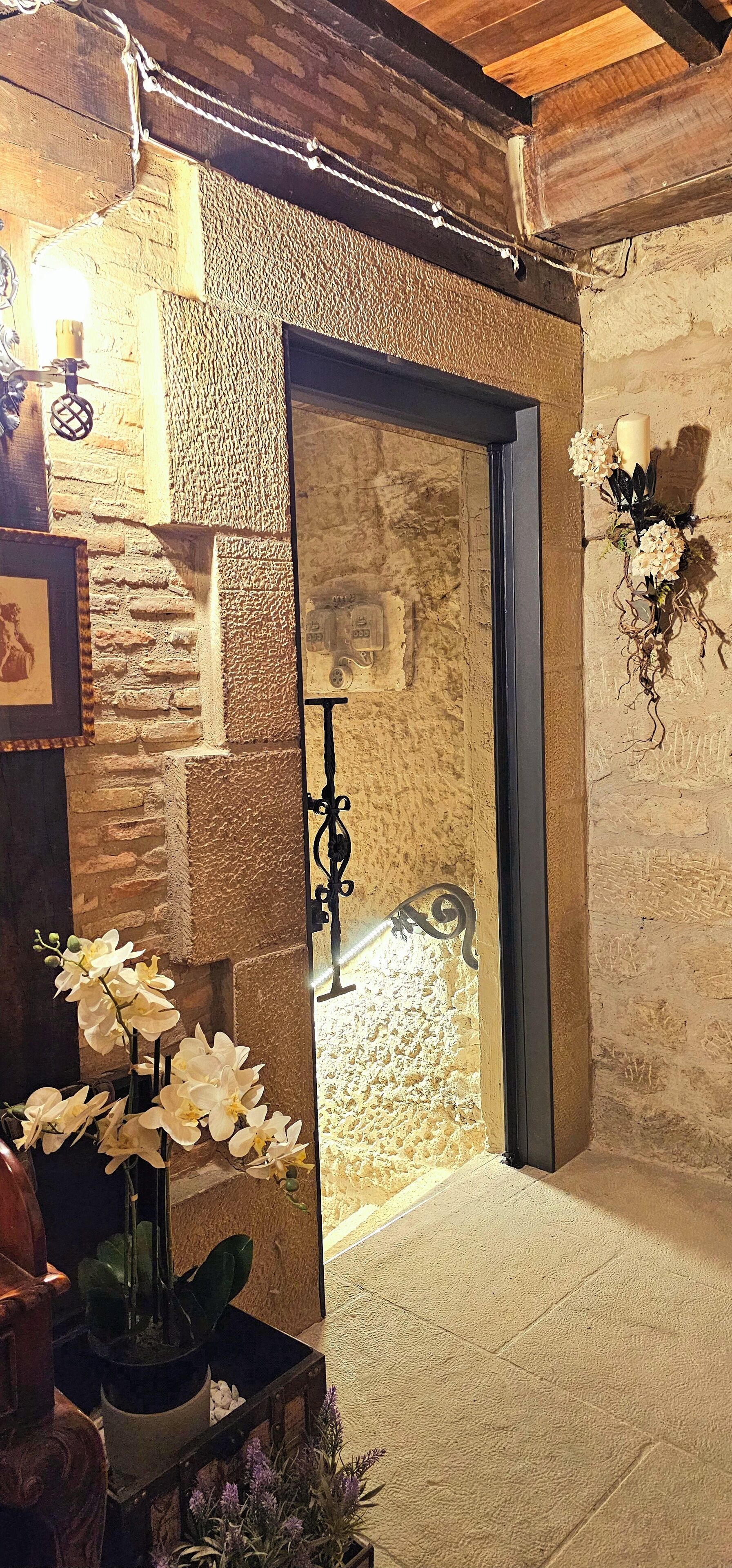 A rustic interior with a stone wall, a wooden door, and a small staircase leading to an upper level. There are potted plants and a candle on the wall.