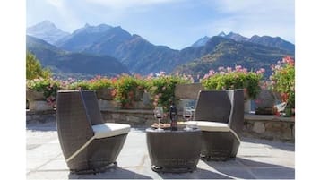 A terrace with stone tables and chairs, surrounded by flowers and a mountain view.
