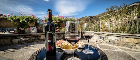 Una mesa con una botella de vino, dos copas de vino y un bol de comida en un patio de piedra con vista a un paisaje montañoso.