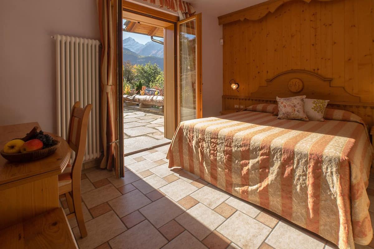 Un dormitorio con cabecera de madera, cama con ropa de cama a rayas, escritorio de madera con frutero, y vistas a montañas a través de un ventanal abierto.