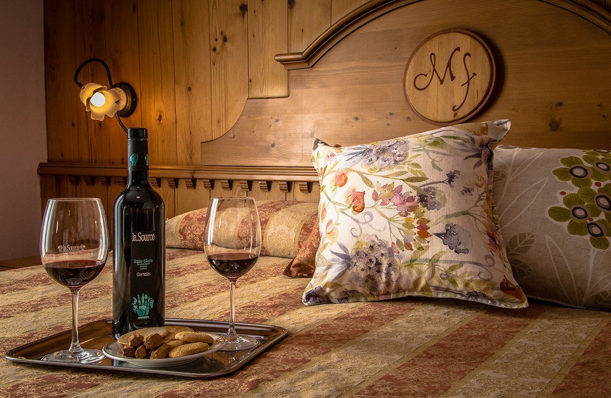 A wooden bed with floral pillows, a bottle of wine, two wine glasses, and a tray with cookies.
