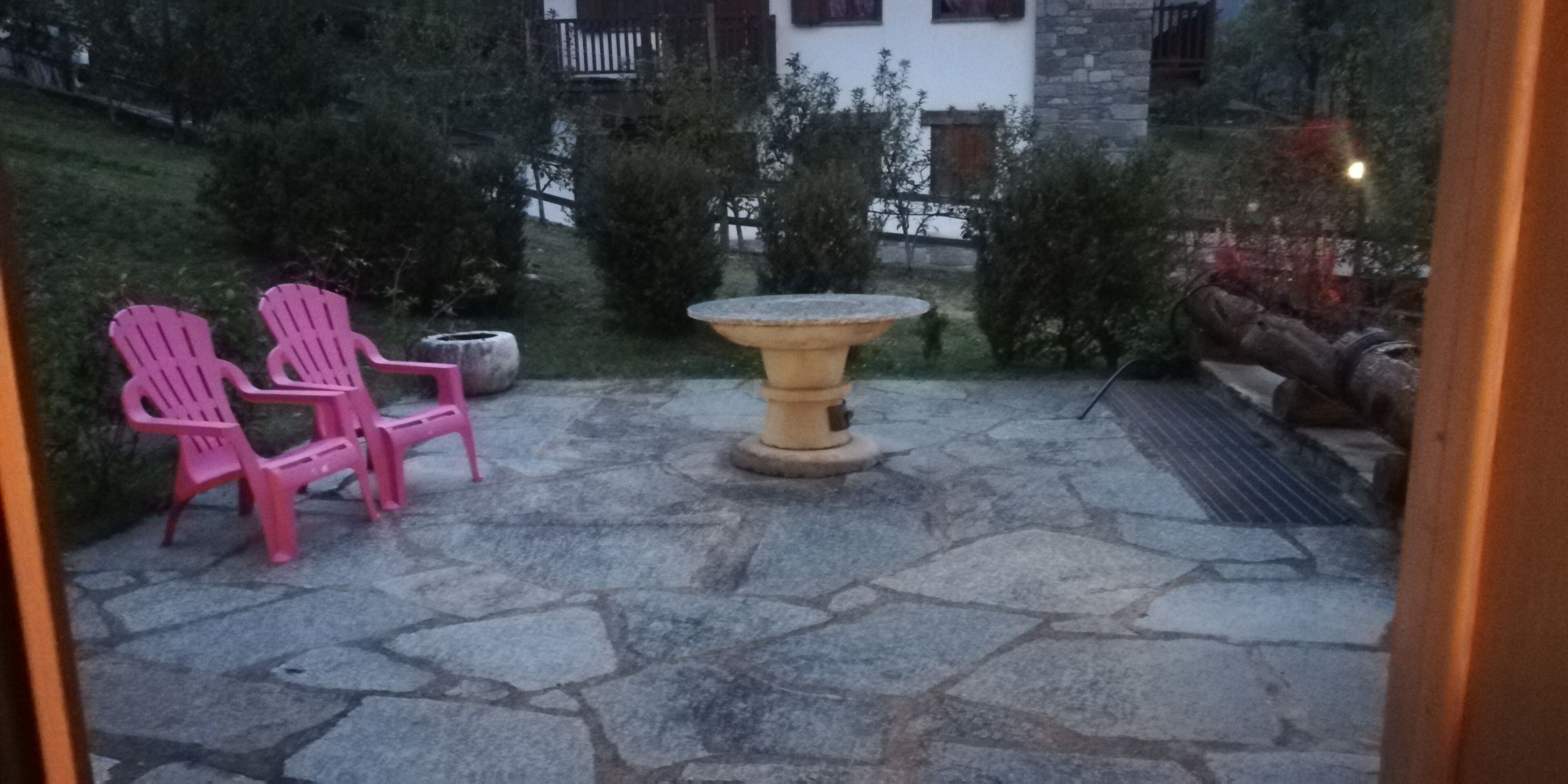 En uteplats med stenbeläggning, två rosa stolar, ett runt bord och en blomkruka i sten.