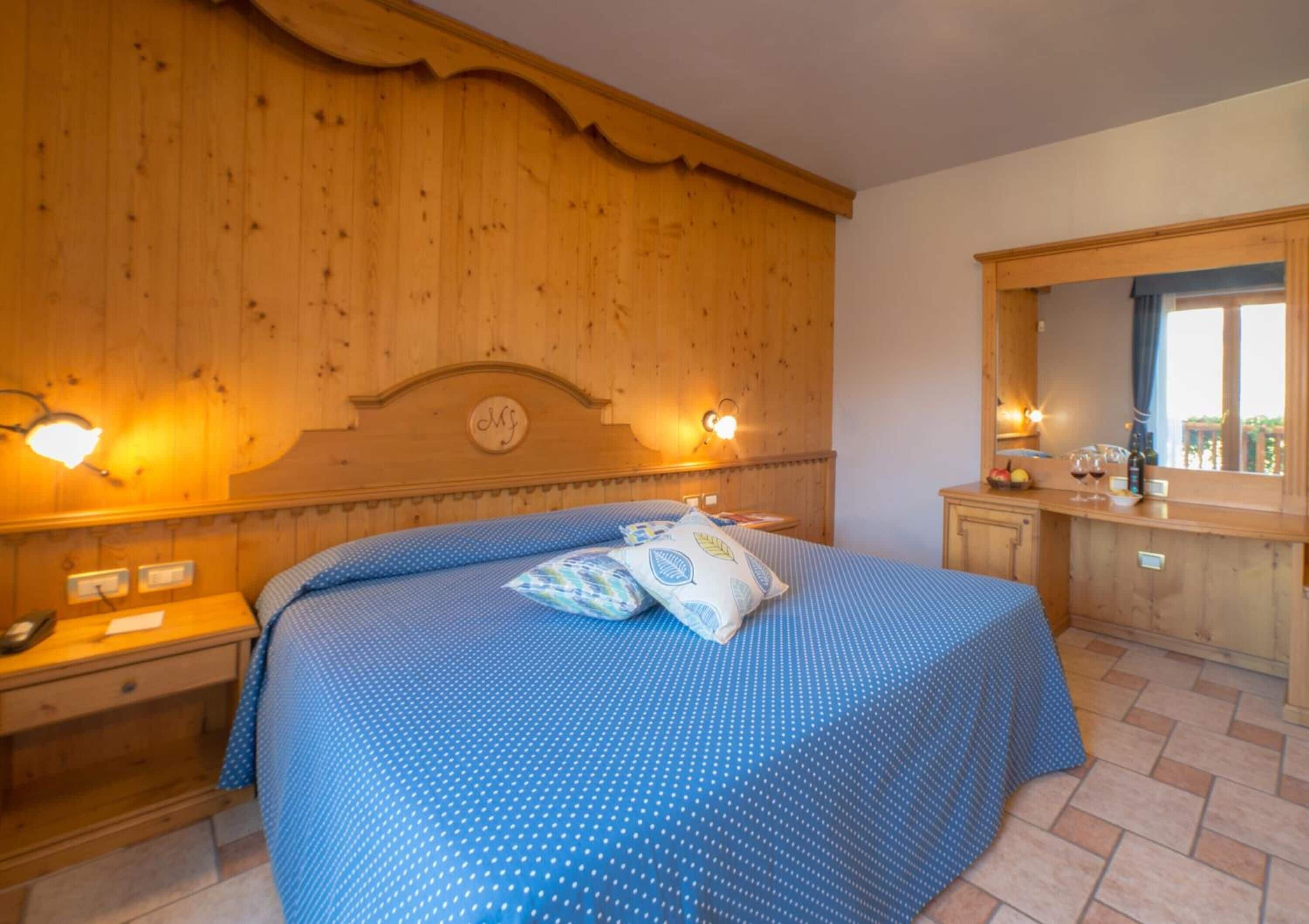 A bedroom with a wooden headboard, a large bed with blue bedding, two bedside tables, a mirror, and a small window.