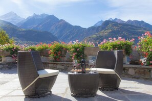 Two wicker chairs with cushions, a bottle of wine, and glasses on a table, set against a mountainous backdrop with flowering plants. - Hotel Lo Fleye (Saint-Pierre)