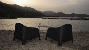 Een tafel met twee stoelen op een kiezelstrand, met uitzicht op een rustig waterlichaam en bergen op de achtergrond.