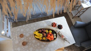 Een eettafel met een fruitmand en schalen vol fruit, een rieten stoel en een klein tafeltje met een kopje en schotel.