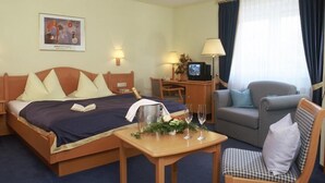 Ein Hotelzimmer mit einem Bett, einem Schreibtisch mit Fernseher, einem kleinen Tisch mit Pflanze und Gläsern, einer Couch und einem Sessel.