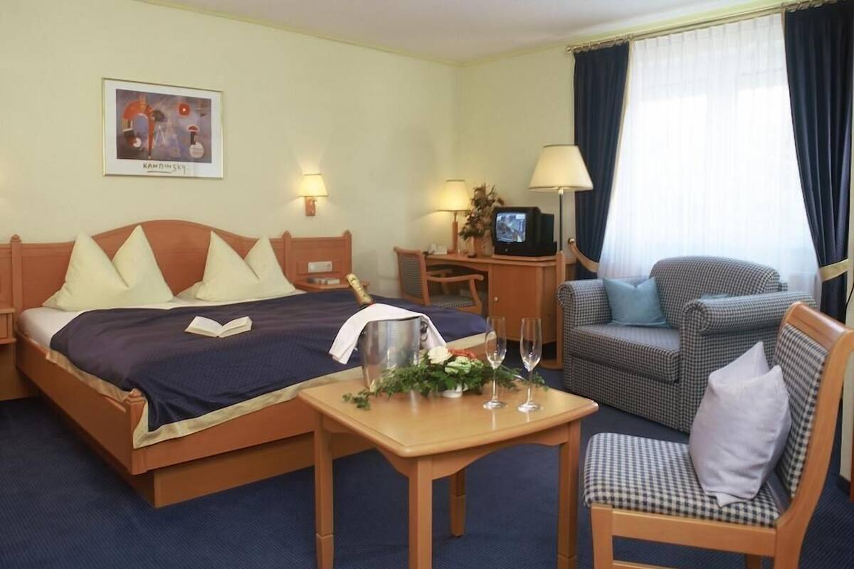 A hotel room with a bed, a desk with a computer, a chair, a small table with a vase, and a window with curtains.