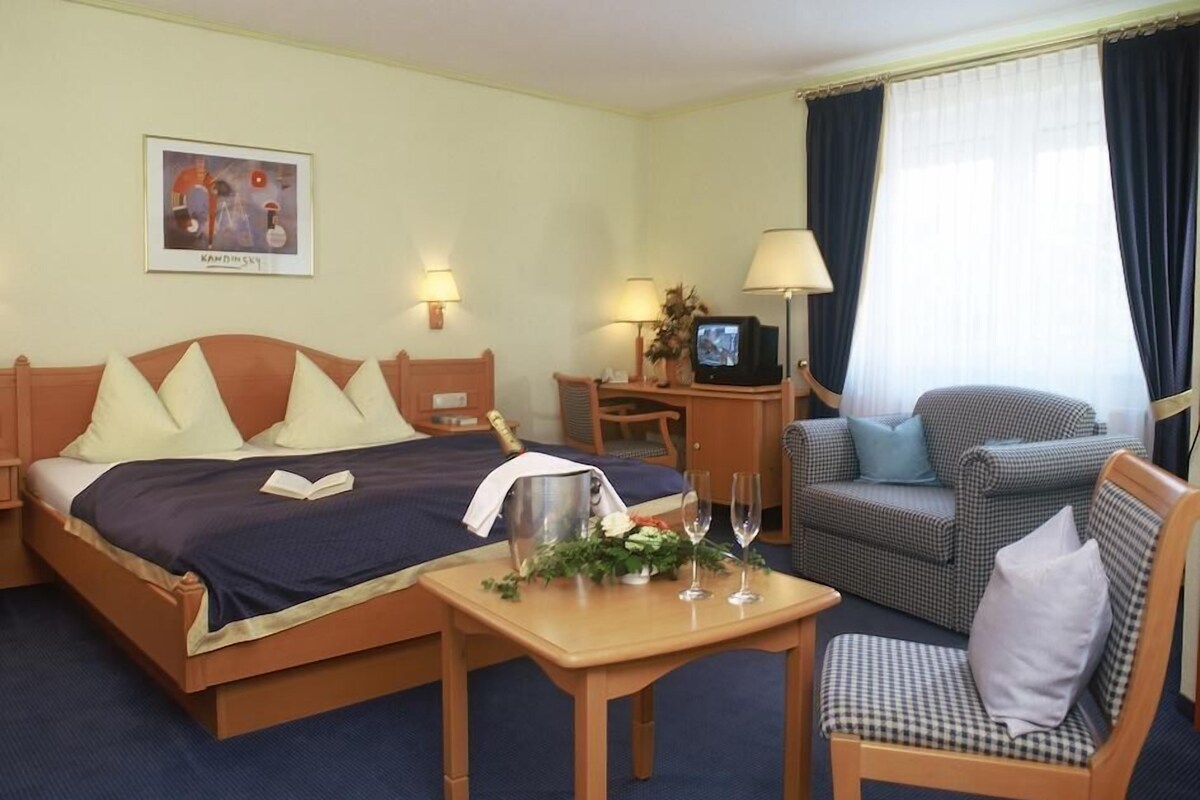 A hotel room with a bed, a desk with a computer, a chair, a small table with a vase, and a window with curtains.