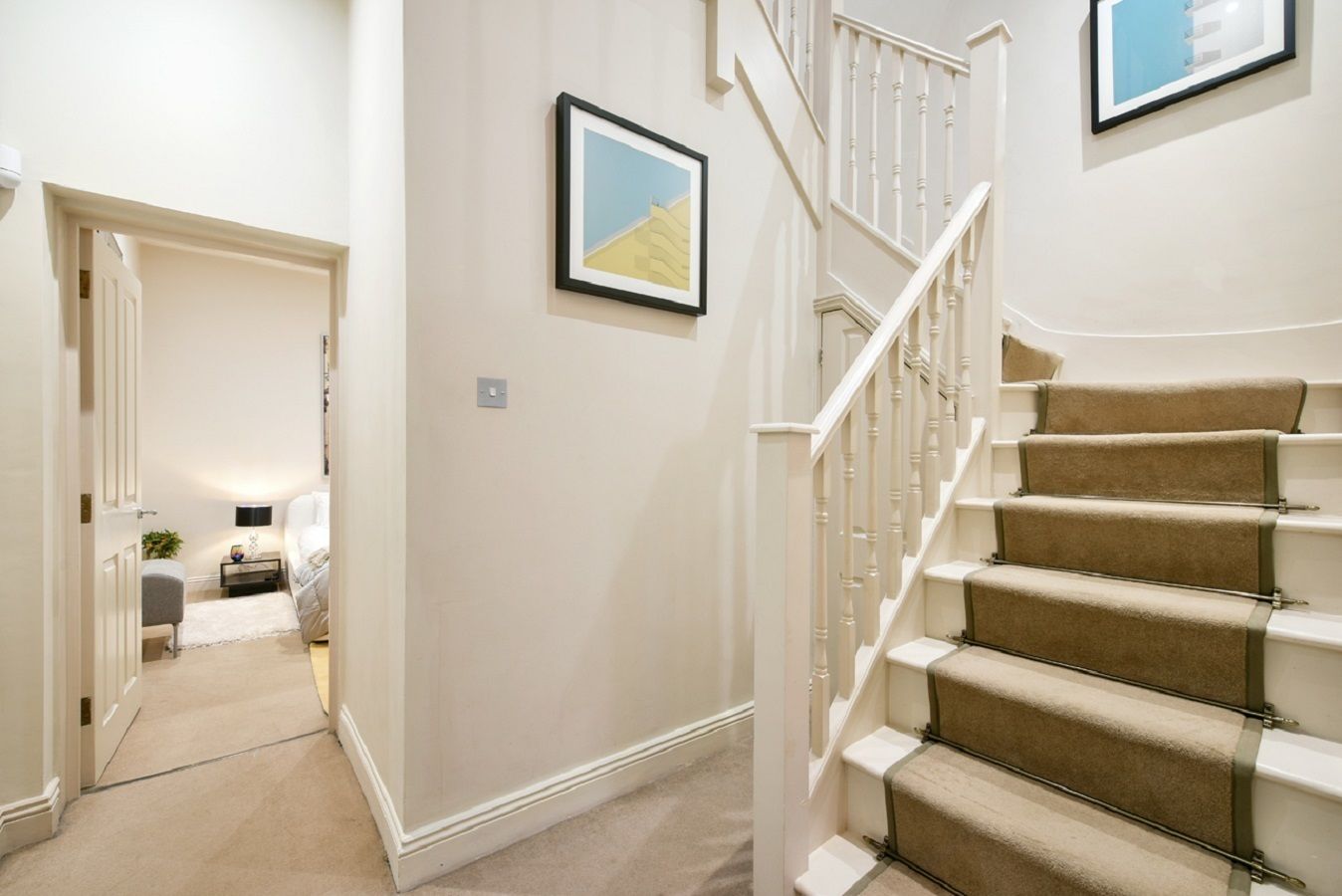 A staircase with beige carpeted steps and white railings, leading to a bedroom with a bed and bedside table.