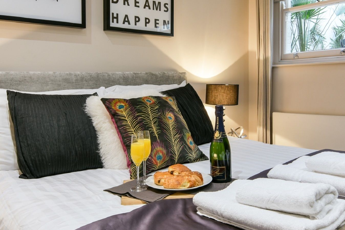 A neatly made bed with a pillow, a bottle of champagne, a plate with a pastry, and a glass of orange juice.