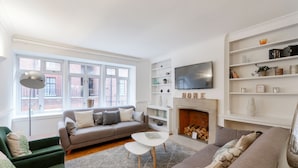 A modern living room with a grey sofa, a green armchair, and a white coffee table. There is a fireplace with a mantel and a TV mounted above it. - Cavendish - By Sojourn (London)