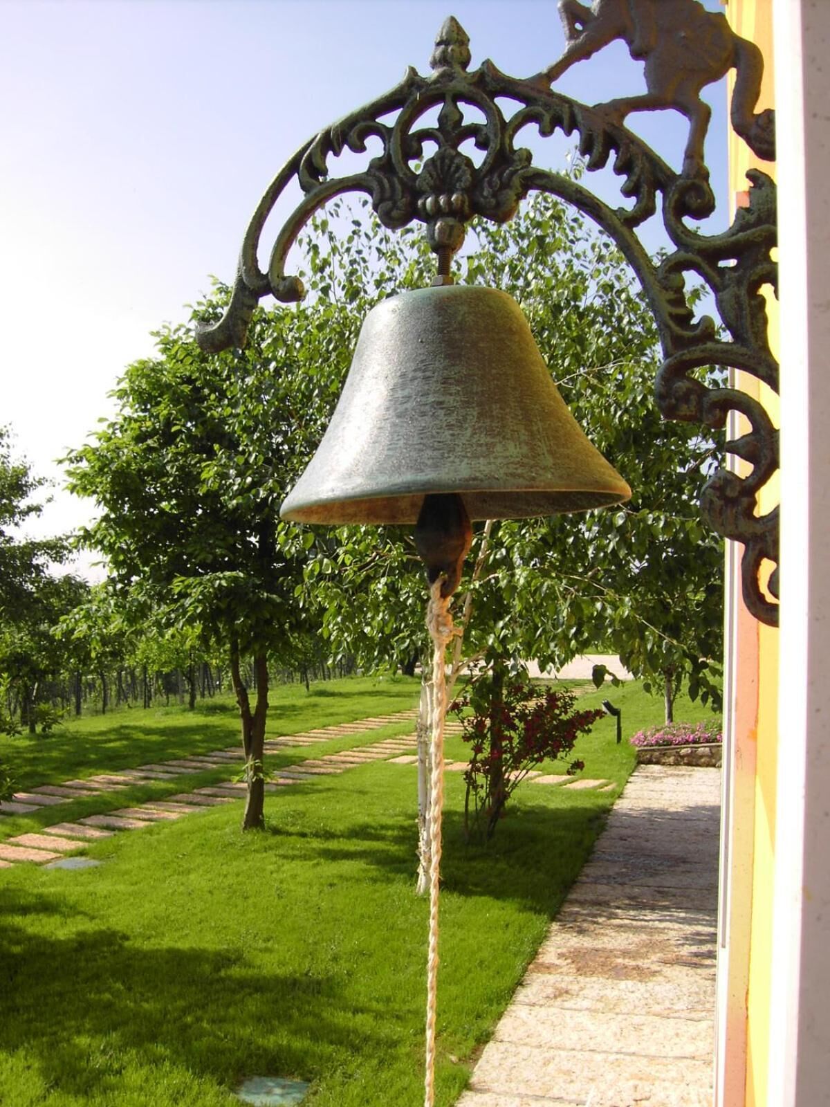 Un jardín con una campana metálica grande montada sobre un poste decorativo, rodeado de abundante follaje y un sendero de piedra.