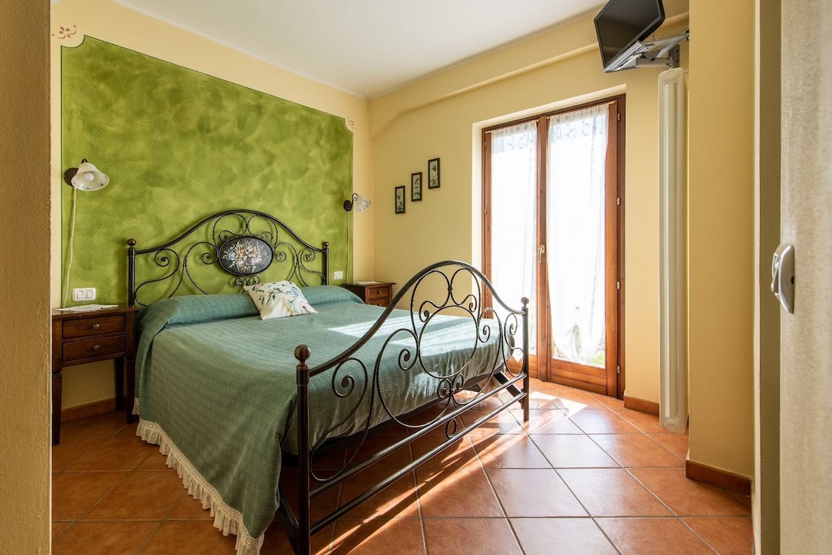 A bedroom with a green wall, a metal bed frame, a bedside table, a nightstand, a window with a view, and a door.