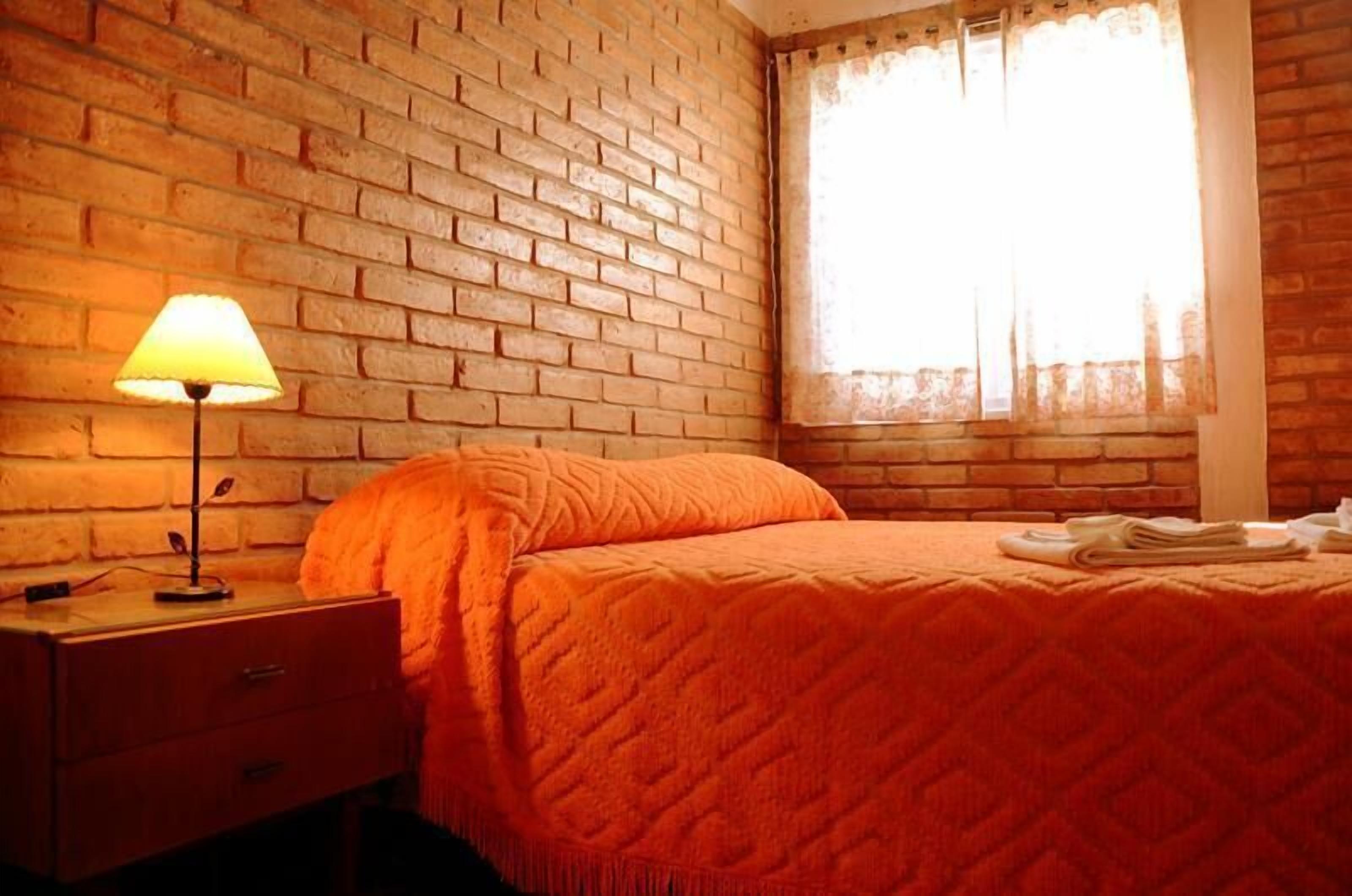 A room with a brick wall, a bed with an orange quilt, a nightstand with a lamp, and a window with sheer curtains.