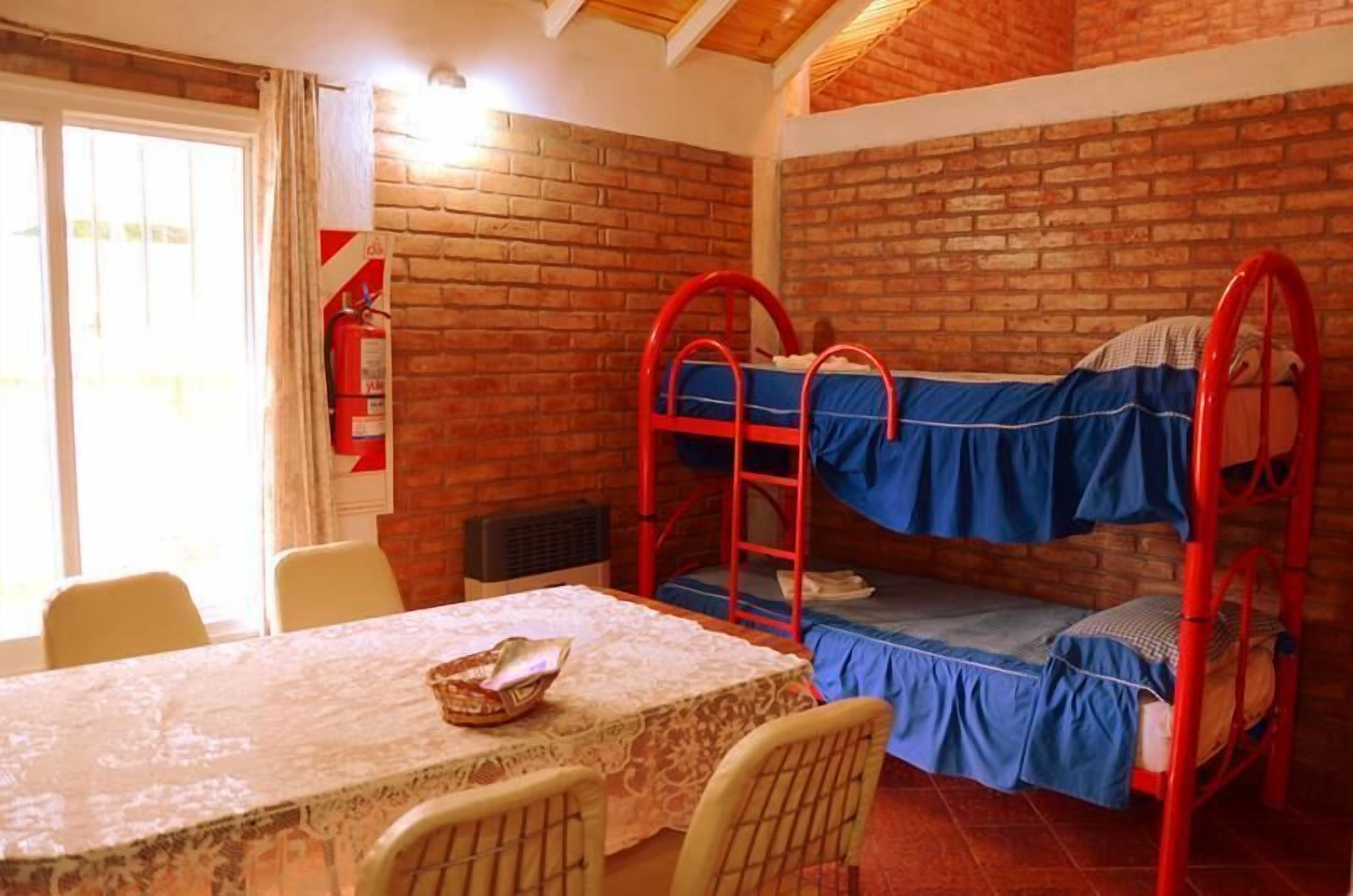 Una habitación con pared de ladrillo, una cama litera, una mesa con mantel de encaje y una ventana con cortinas.