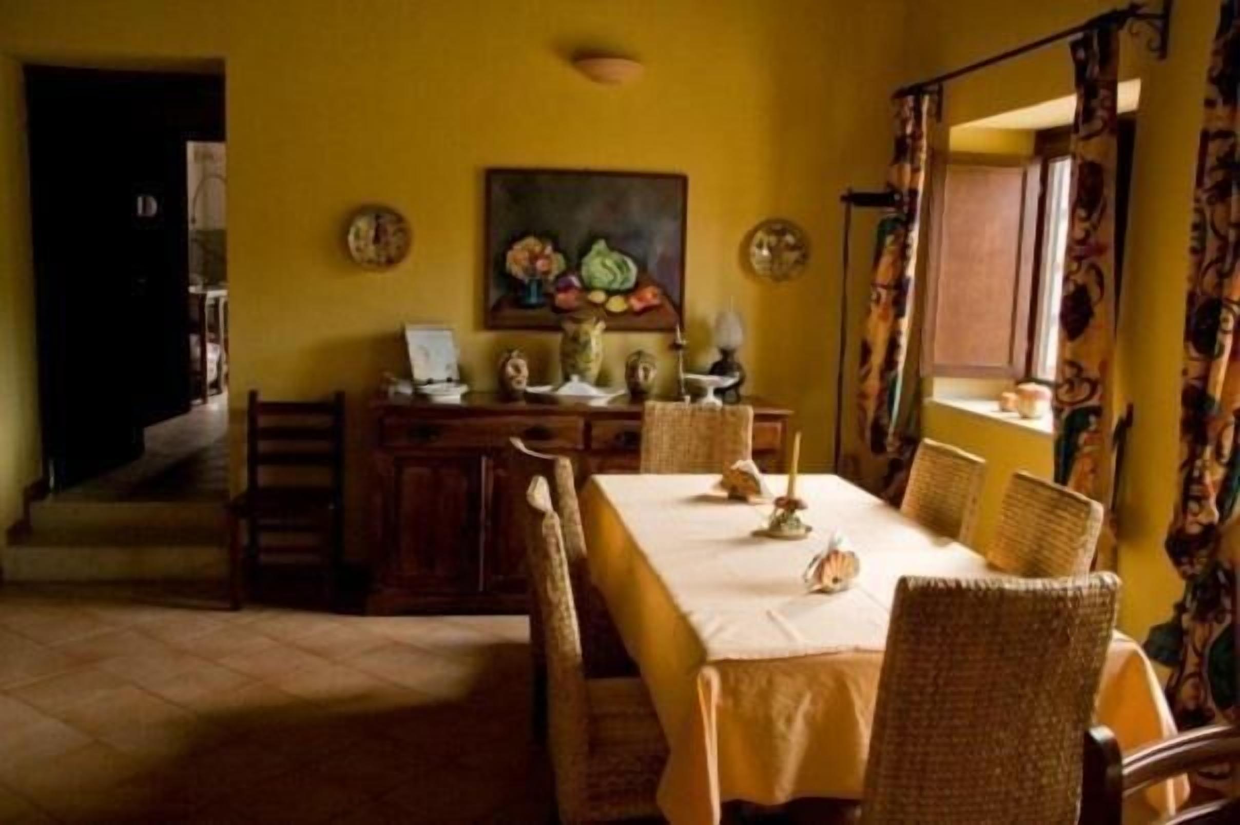 Un comedor con una mesa dispuesta para cuatro personas, rodeada de sillas de mimbre. Un cuadro de frutas cuelga en la pared sobre la mesa.