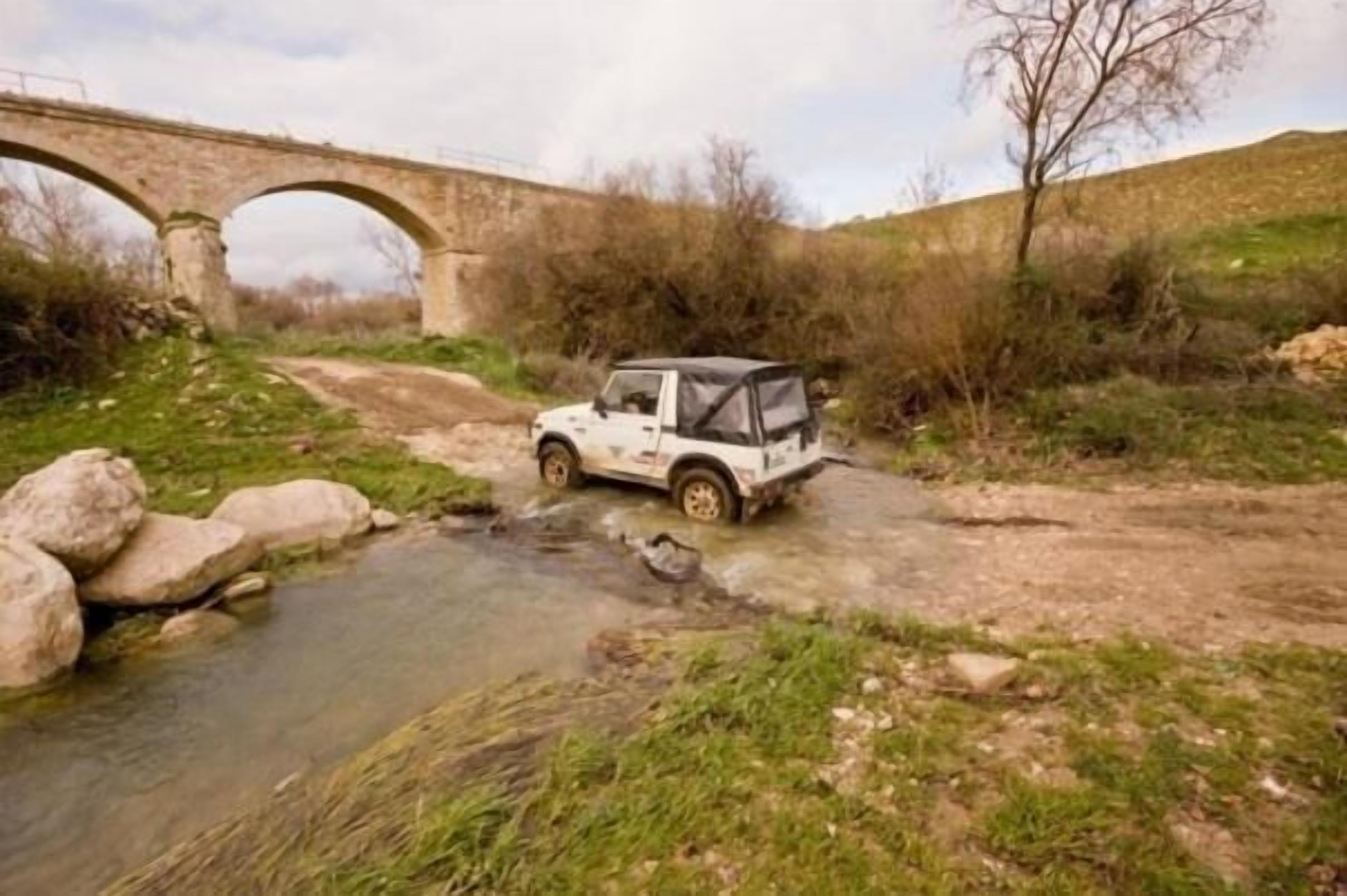 A white off-road vehicle parked near a stream with a stone bridge in the background.