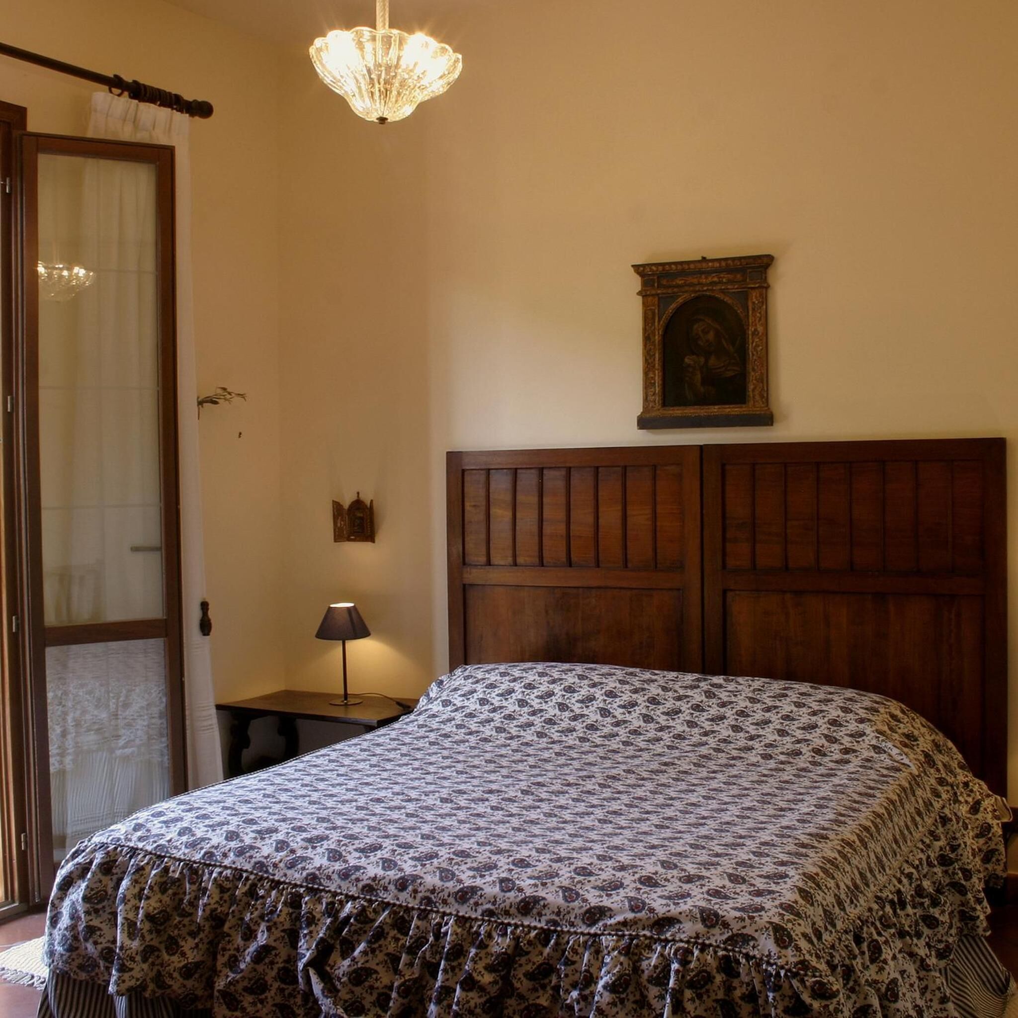 Une chambre à coucher avec une tête de lit en bois, une couvre-lit à motifs, une table de chevet avec une lampe, un tableau encadré au mur et une porte avec un rideau.