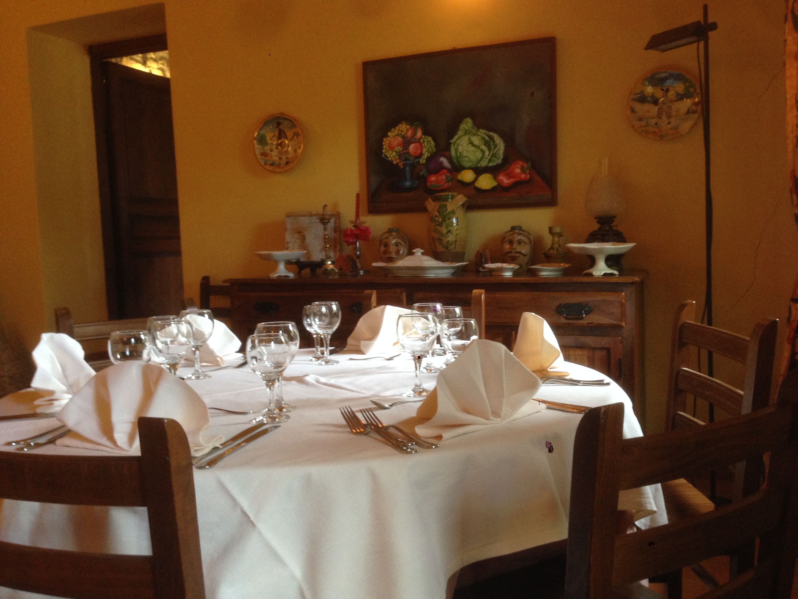 Une table à manger est dressée avec une nappe blanche, des assiettes, des verres et des couverts. Des chaises sont disposées autour de la table, et un tableau représentant des fruits est accroché au mur.