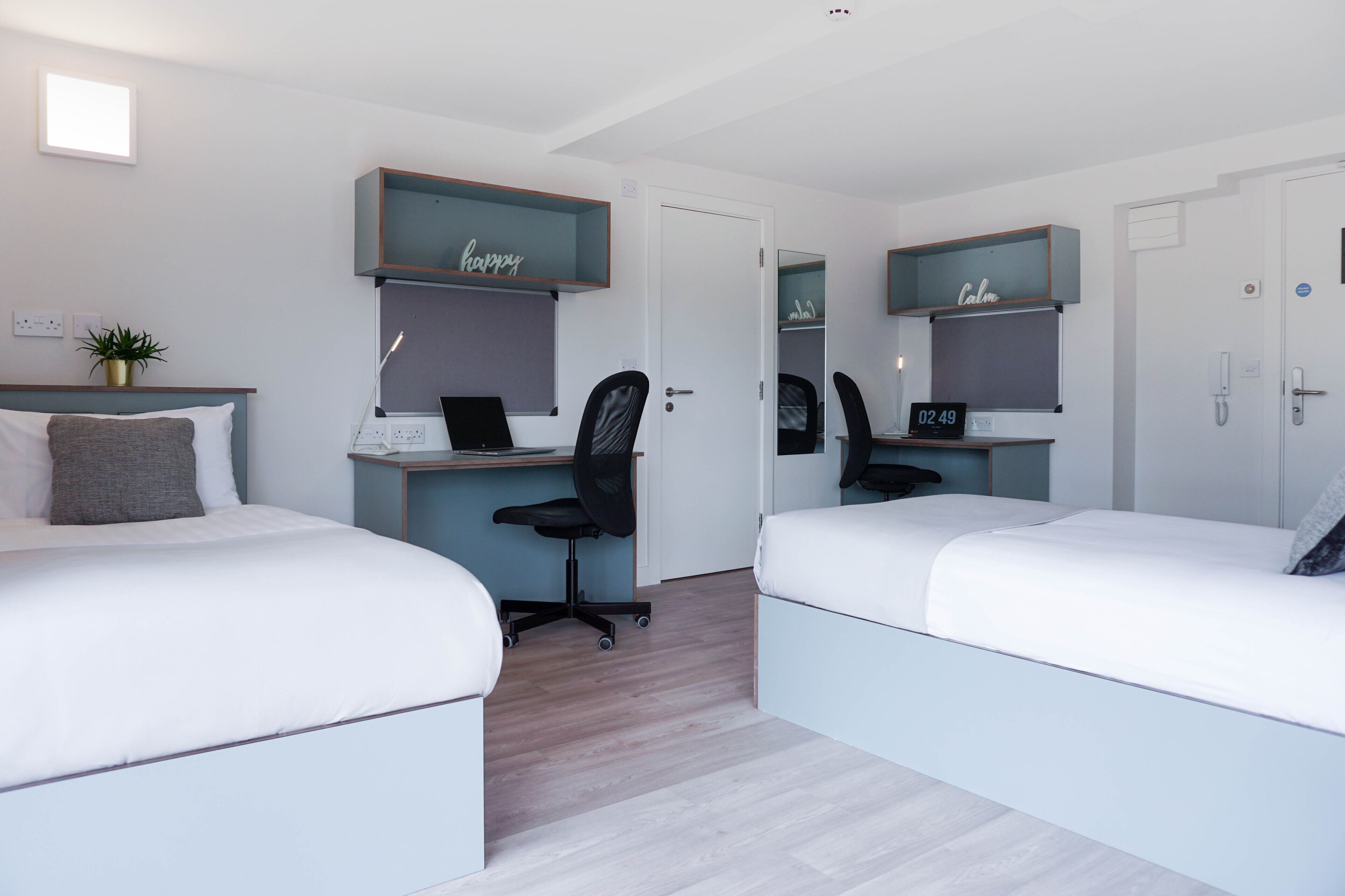 A modern bedroom with two beds, a desk, and a chair. There are shelves above the desk with decorative items.