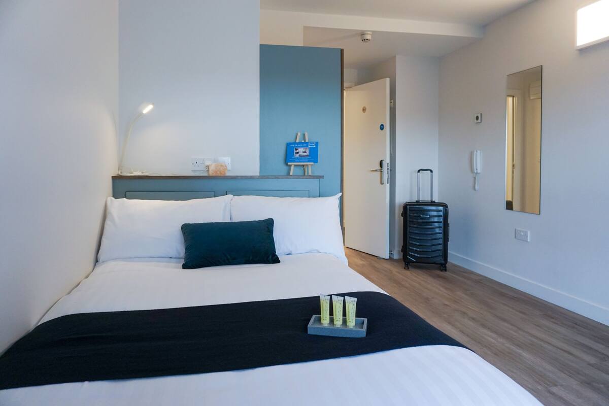 A hotel room with a bed, a bedside table, a lamp, a mirror, a suitcase, and a sign on the wall.