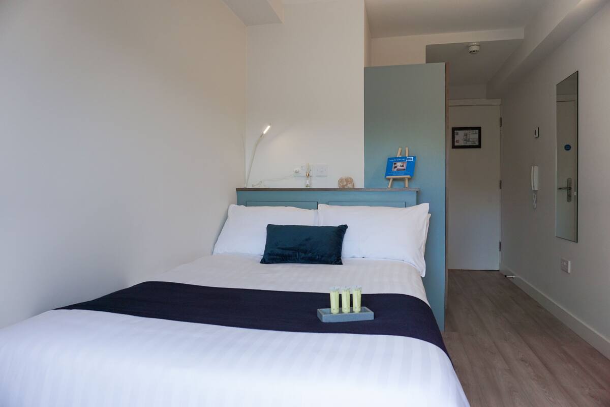 A neatly made bed with a dark blue headboard, a bedside table with a lamp, and a small blue sign on the wall.