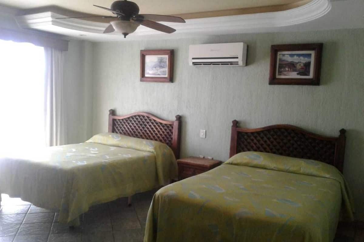 A hotel room with two single beds, a ceiling fan, an air conditioning unit, and framed pictures on the wall.