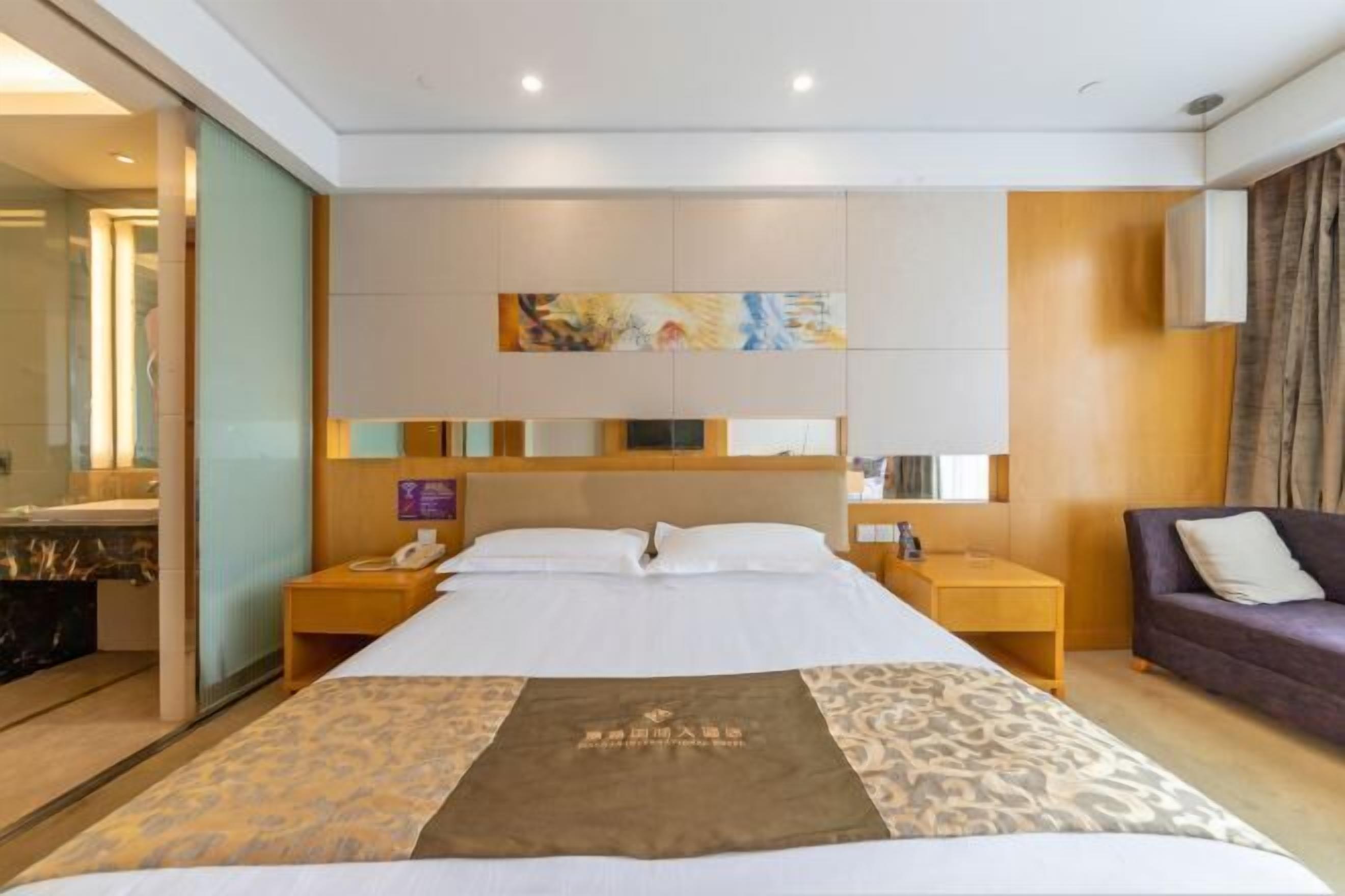 Habitación de hotel con una cama grande, cabecera de madera, mesita de noche con lámpara, una pequeña mesa con teléfono y sofá con cojines.