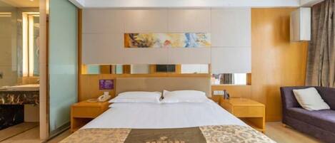 Habitación de hotel con una cama grande, cabecera de madera, mesita de noche con lámpara, una pequeña mesa con teléfono y sofá con cojines.