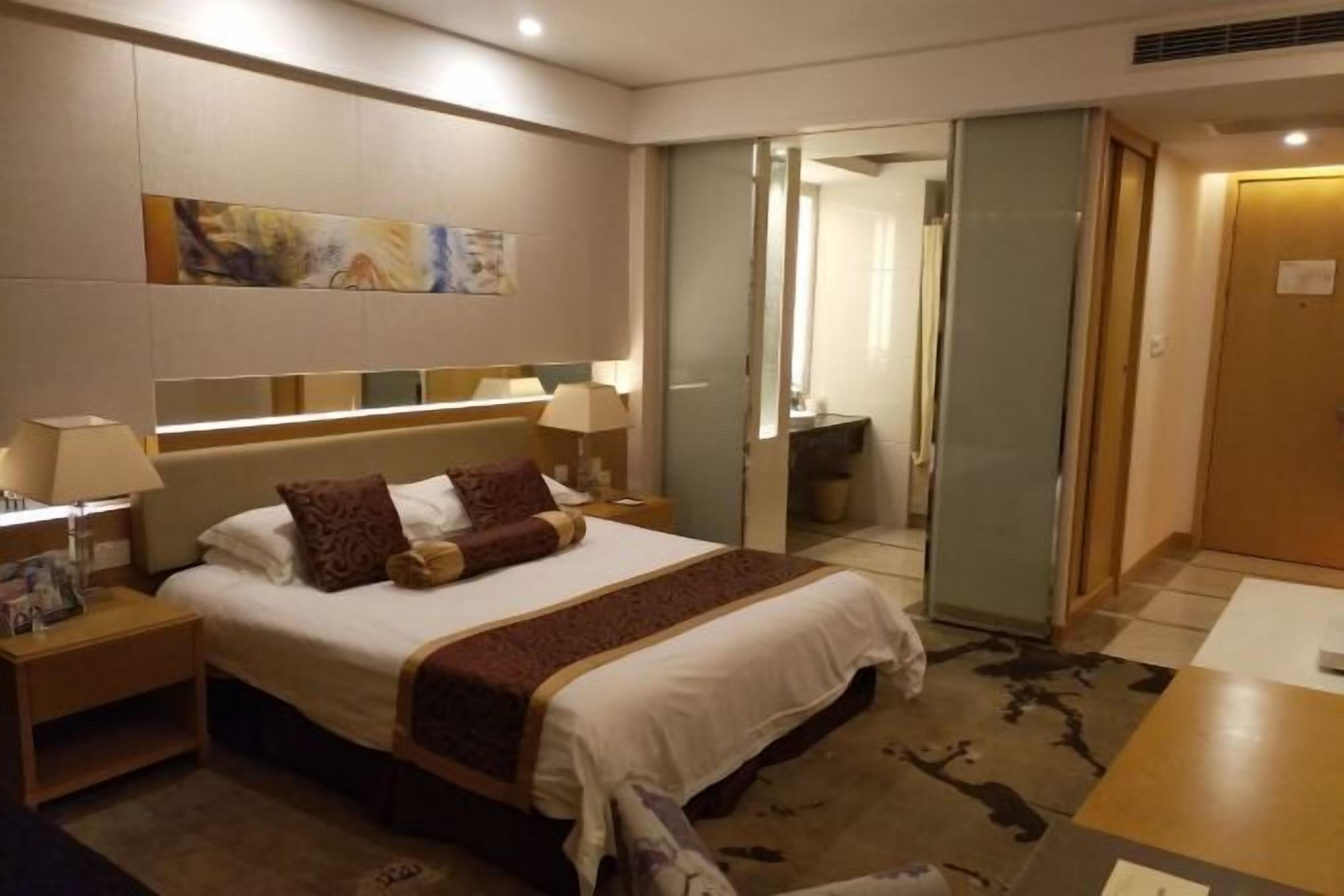 A hotel room with a large bed, bedside tables with lamps, a painting on the wall, and a sliding door leading to a bathroom.
