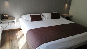 Una cama bien tendida con una colcha blanca y marrón, dos almohadas marrones y una mesita de noche de madera con un teléfono.